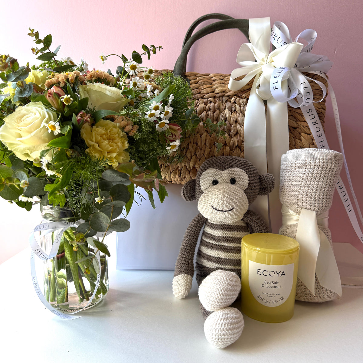Neutral baby gift basket contents with cream and yellow flowers, brown Loui monkey, natural cotton blanket and Ecoya Sea Salt & Coconut candle