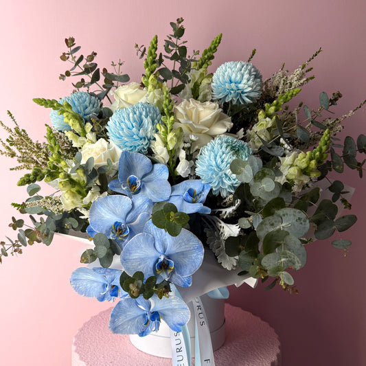Light blue orchids, blue chrysanthemums and white roses arranged with eucalyptus in a white hat box.